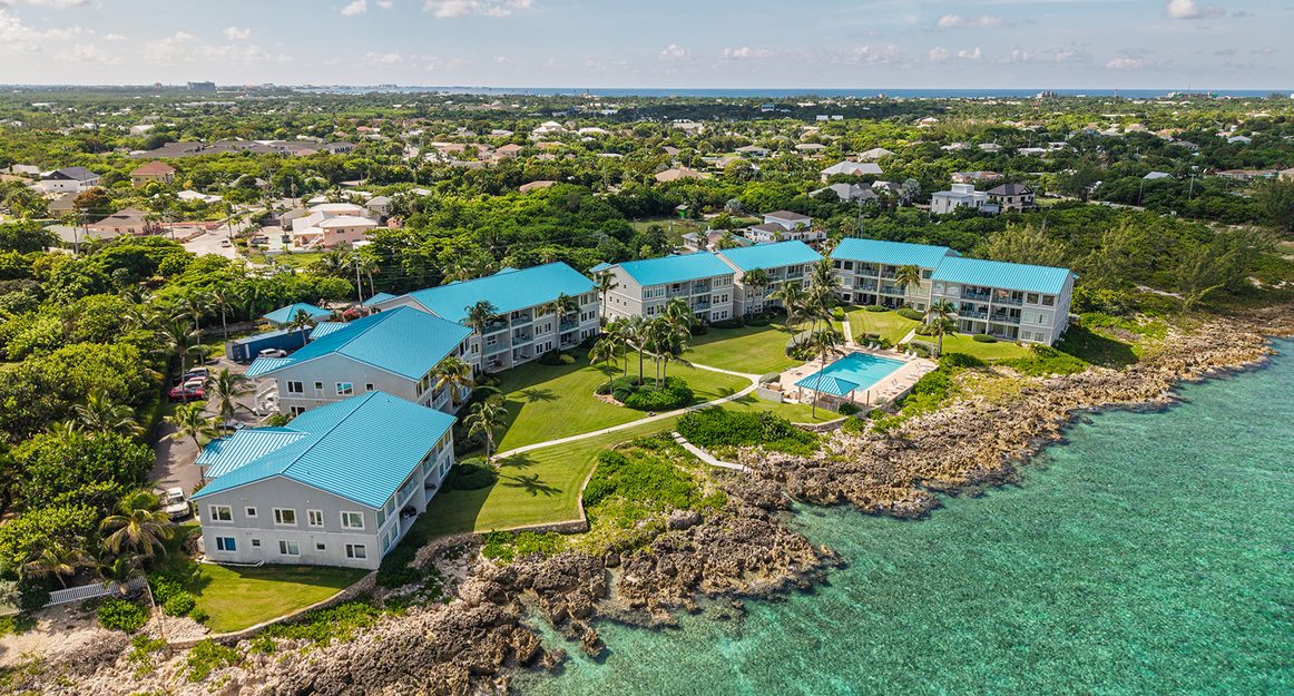 Aerial view of Ocean Pointe Villas on the West Bay ironshore, with the pool set between the blue-roofed blocks and the reef beyond